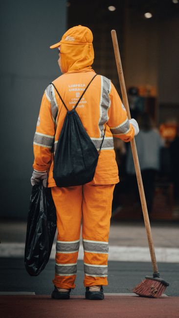 A sanitation worker dressed in a bright green uniform with a matching safety helmet and reflective strips is collecting waste from a black trash bag inside a large green wheeled bin situated on a pave