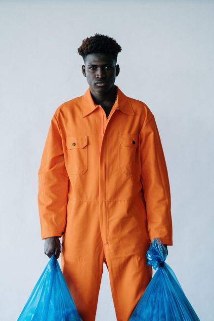 A young male cleaner in an orange jumpsuit holding two blue plastic garbage bags, standing in a well-lit indoor space with a plain light gray background. The surface is clean and smooth, likely a tile