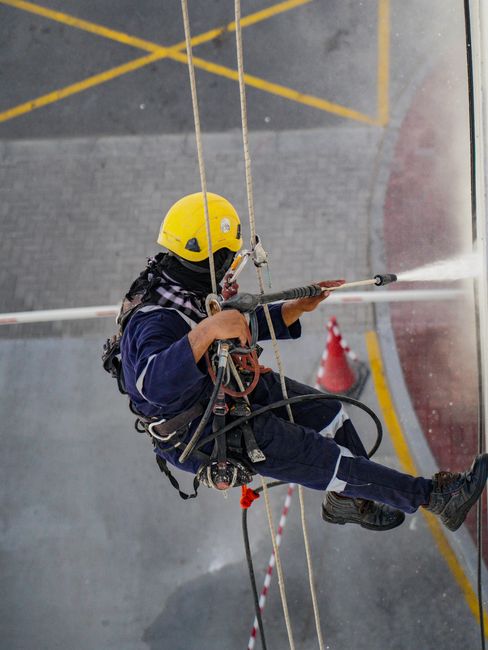 A professional window cleaner from Carpet Cleaners SE10 is photographed mid-action, suspended on a harness while cleaning large glass panels on the exterior of a modern commercial building. The buildi